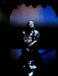 Pan Daijing sitting in dramatic low-key lighting wearing a white collared shirt reflected on a dark glossy surface.