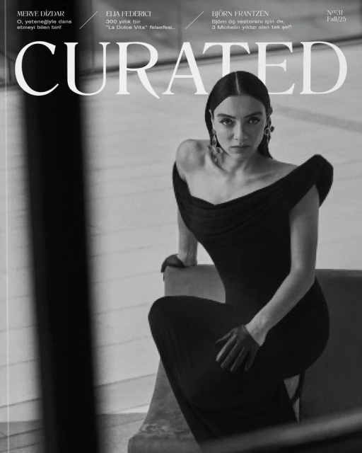 A monochrome fashion magazine cover with a woman in a black dress, striking a seated pose, featuring a curated layout and bold typography.