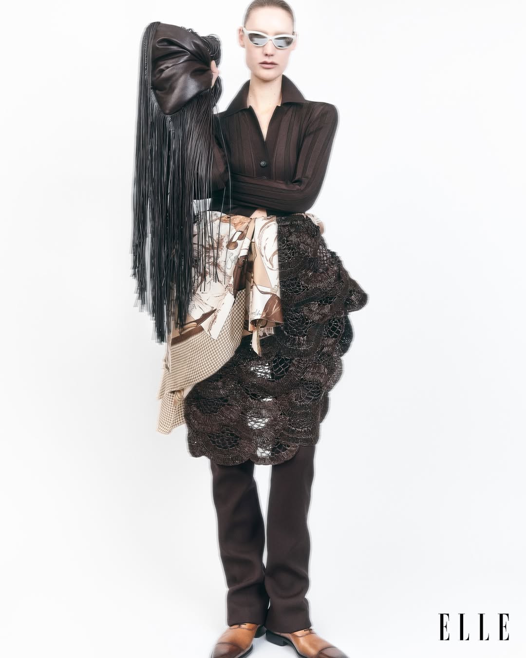 Fashion editorial shot featuring a female model in an unconventional outfit with a brown top, layered skirts, and sunglasses against a white backdrop.