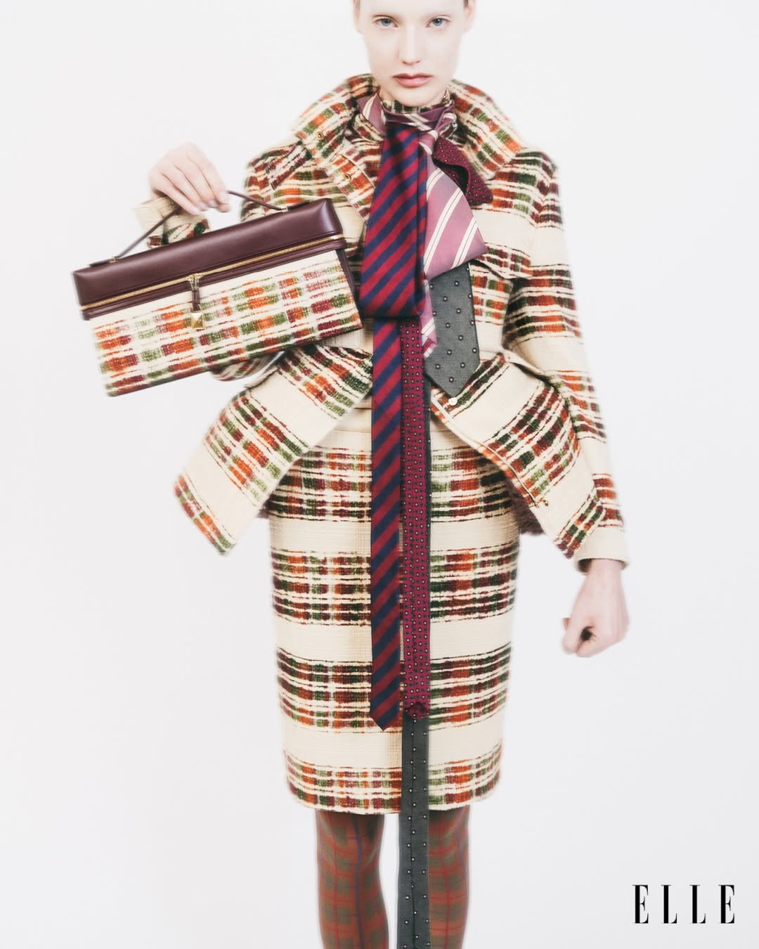 Fashion editorial shot of a fair-skinned woman wearing a plaid skirt suit, three ties, and holding a structured purse against a white background.