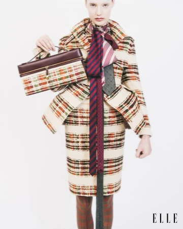 Fashion editorial shot of a fair-skinned woman wearing a plaid skirt suit, three ties, and holding a structured purse against a white background.