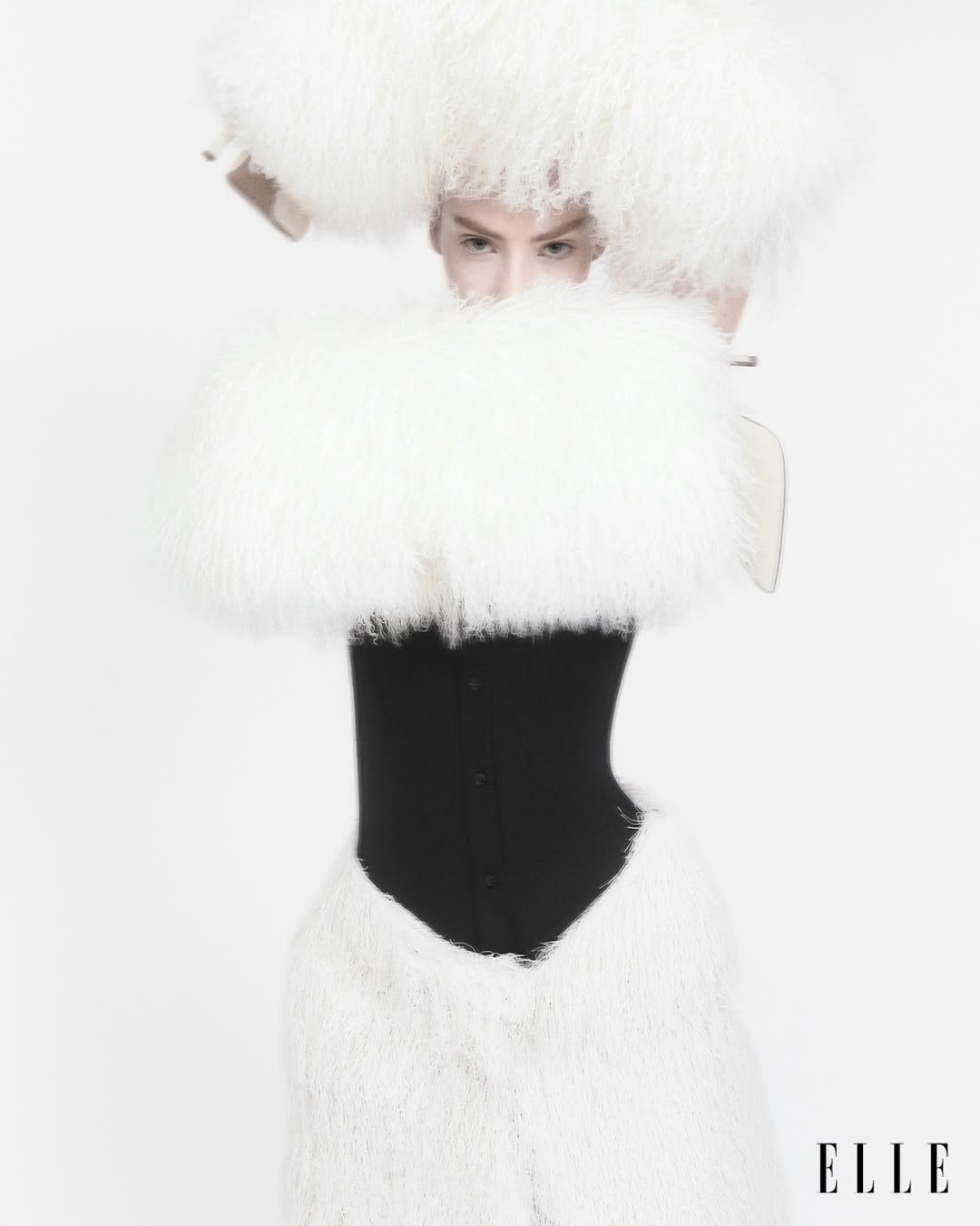 Fashion editorial shot of a model wearing a black corset, white furry pants, and a large furry white headpiece on a white background.