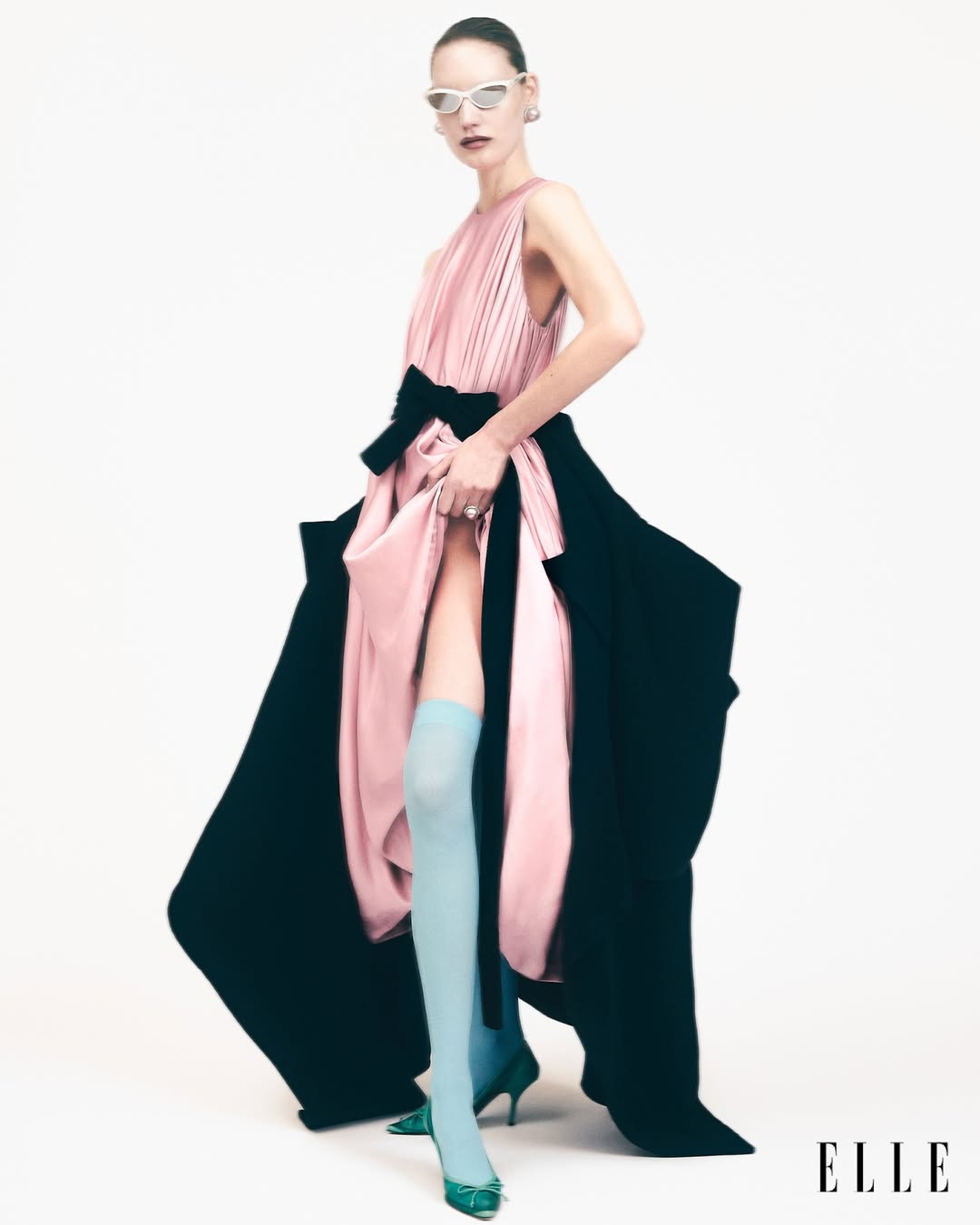 A model poses in a pink gown with a black sash and black side panels, accessorized with light blue socks and white sunglasses.