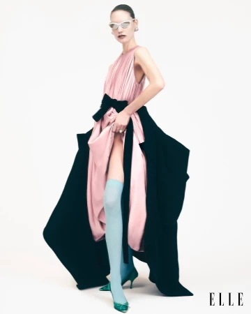 A model poses in a pink gown with a black sash and black side panels, accessorized with light blue socks and white sunglasses.
