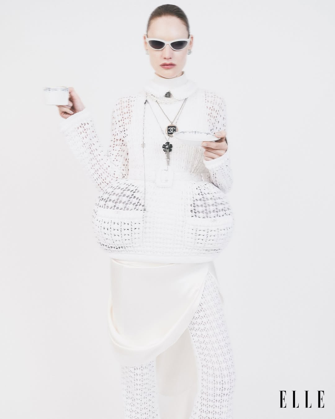 A woman in all-white fashion stands in a photo studio, holding a tea cup and saucer. She wears sunglasses, jewelry, and knitted fabric clothing.