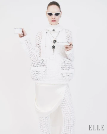 A woman in all-white fashion stands in a photo studio, holding a tea cup and saucer. She wears sunglasses, jewelry, and knitted fabric clothing.