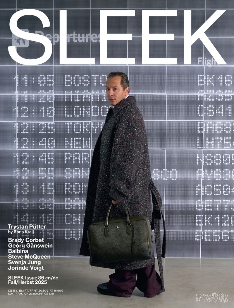 Man on cover of Sleek magazine in gray coat with airport display in the background. He's holding a green leather bag.