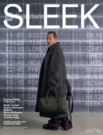Actor Trystan Pütter wearing a long charcoal coat and holding an olive duffle bag in front of an airport departures board.