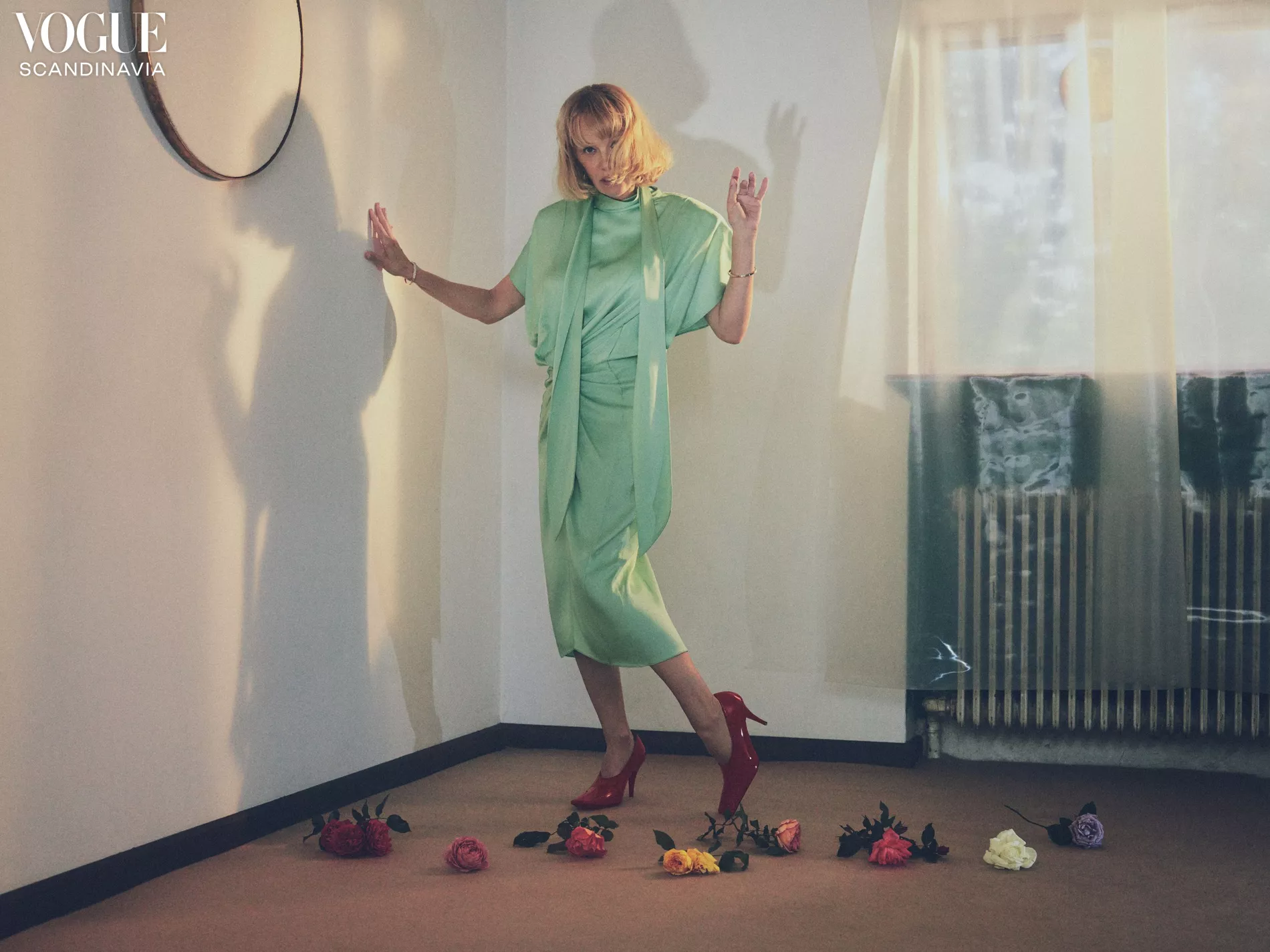 A woman in a green dress and red heels poses in a room with roses on the floor. Vogue Scandinavia logo is visible in the corner.