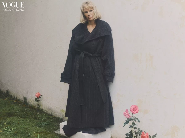 A woman poses in an oversized grey wool coat outdoors, beside a white wall and pale pink roses for a Vogue Scandinavia fashion shoot.