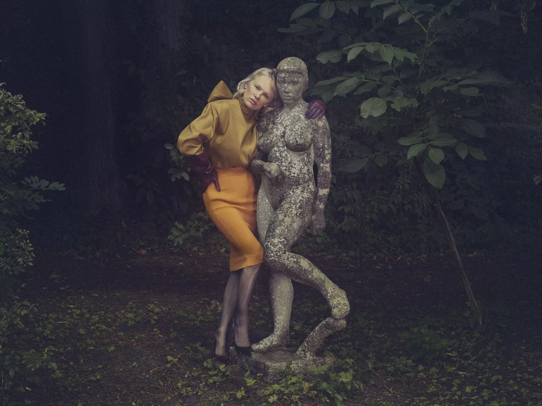 Fashion shot of a woman in mustard blouse and orange pencil skirt leaning on a stone statue in a green outdoor environment.