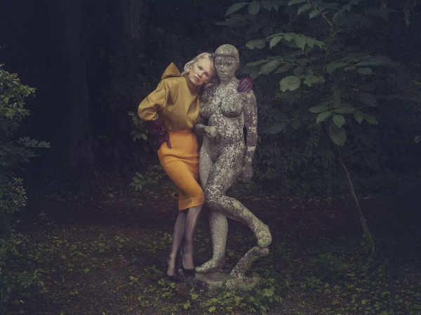 Fashion shot of a woman in mustard blouse and orange pencil skirt leaning on a stone statue in a green outdoor environment.