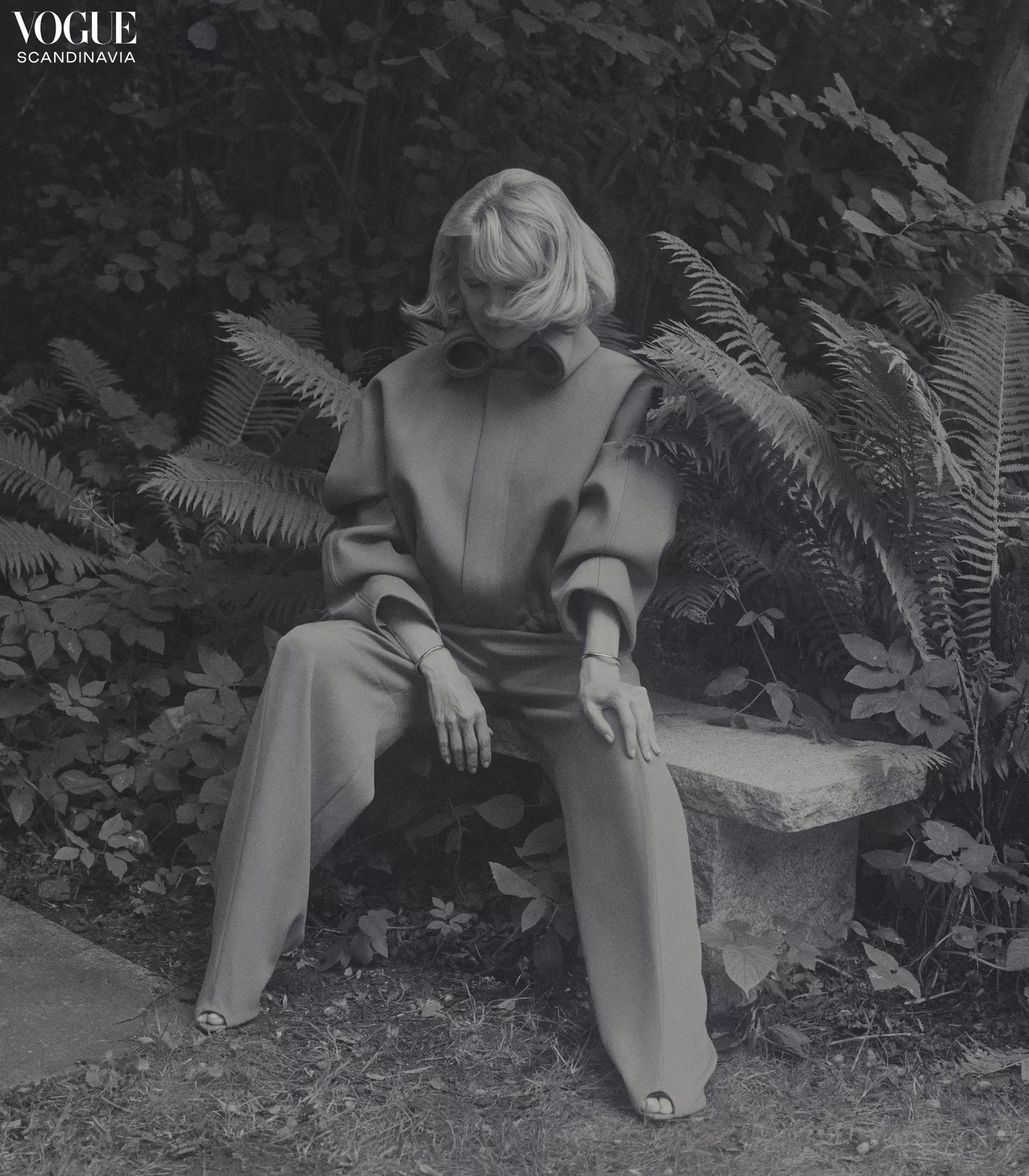 Monochromatic fashion shot: Woman in tailored clothing seated on a stone bench surrounded by lush foliage, evoking a vintage editorial feel.