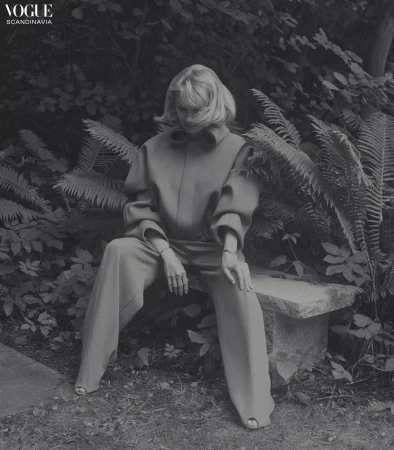 Monochromatic fashion shot: Woman in tailored clothing seated on a stone bench surrounded by lush foliage, evoking a vintage editorial feel.