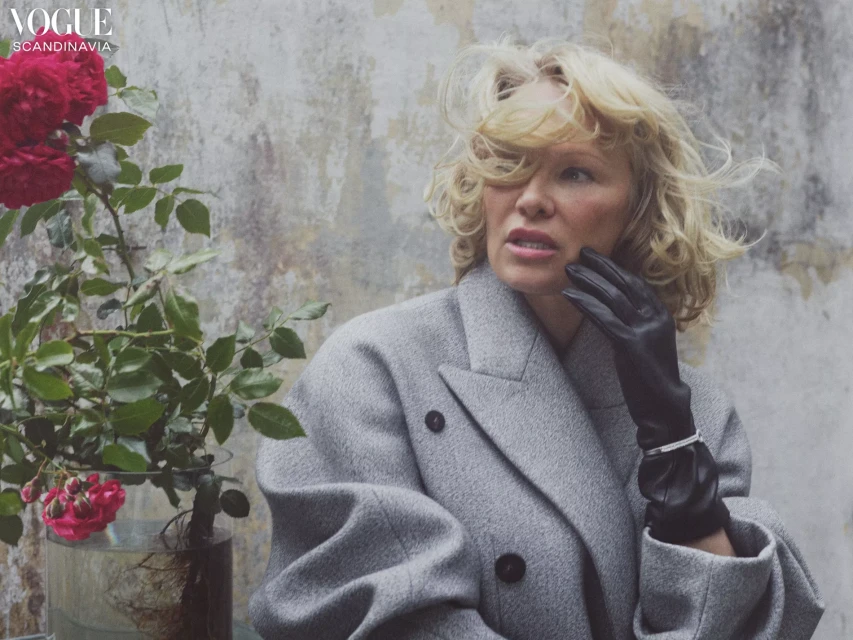 Woman in a grey coat and leather gloves poses next to a glass vase filled with red roses and green leaves against a textured wall.