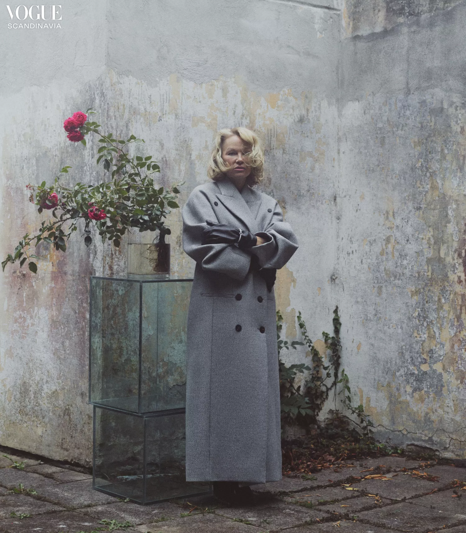 Fashion photography featuring a blonde woman in a grey coat next to glass cubes and a rose bush against a textured wall outdoors.