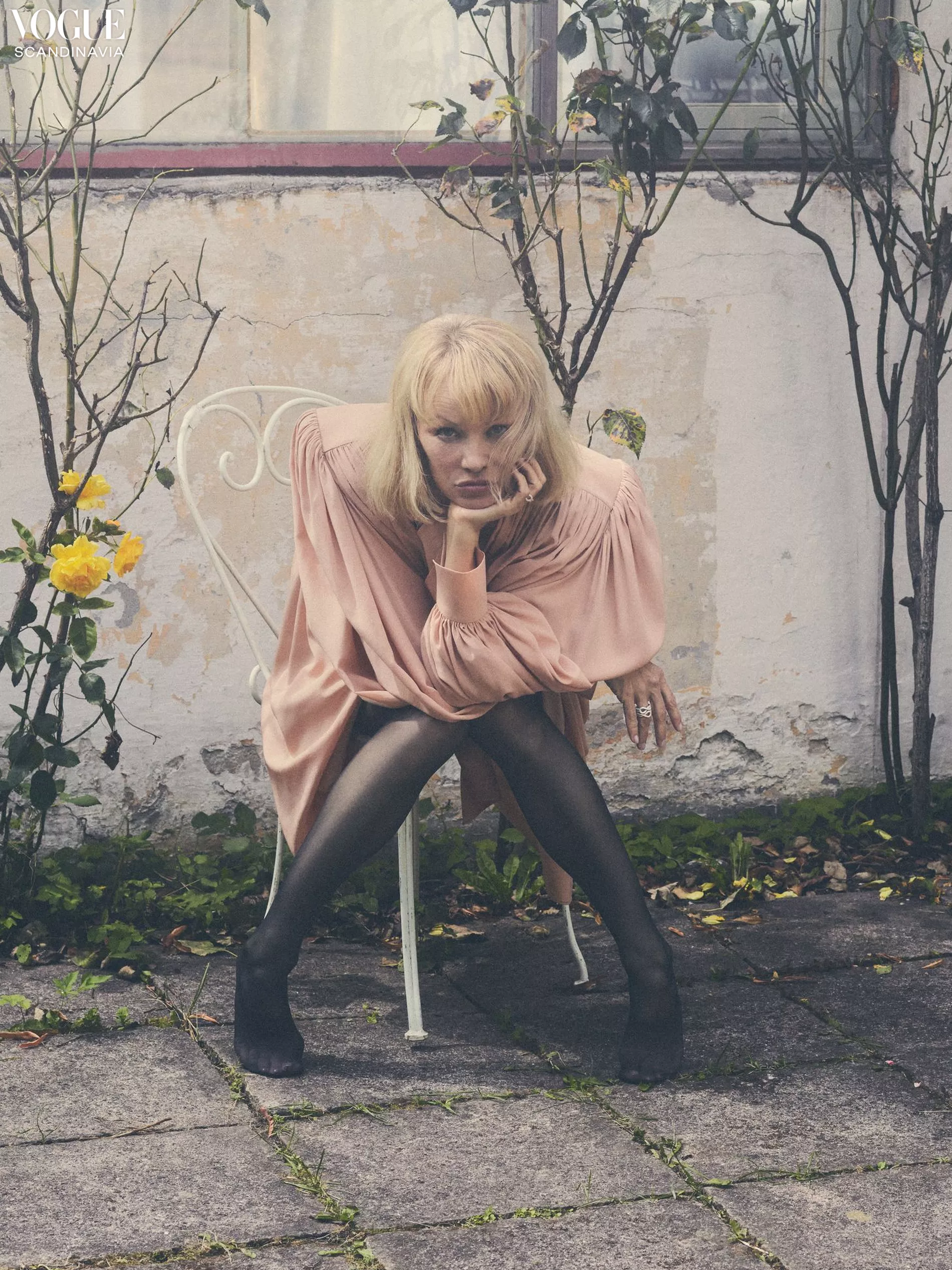 A woman with blonde hair in a pink dress and black tights sits on a white chair outside near a building with trees and yellow flowers.