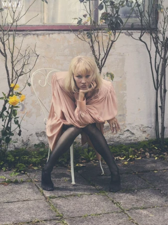 Blonde woman wearing a pink draped dress and sheer black stockings seated on a white wire chair outdoors.
