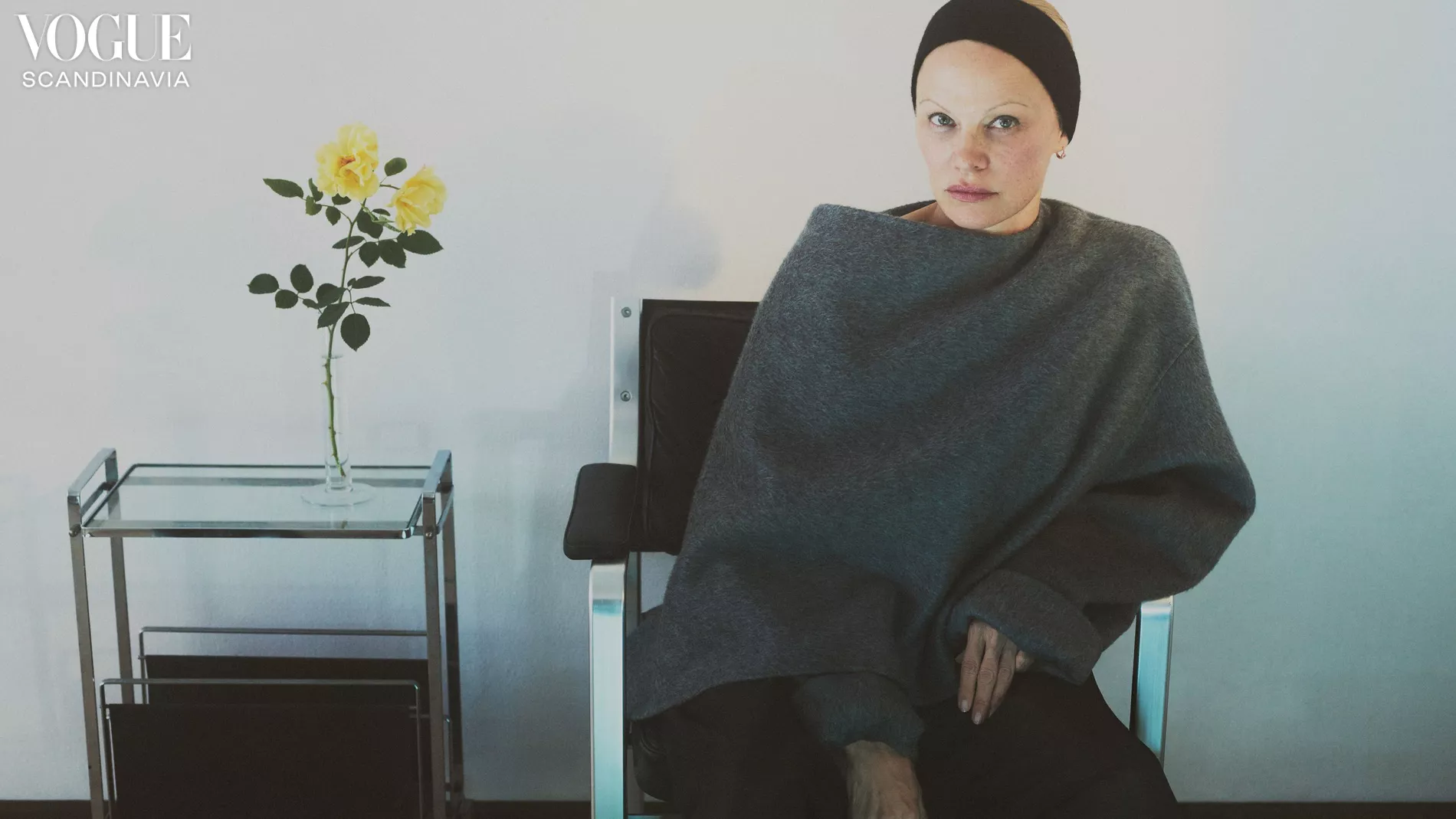 Portrait of adult female model with headband, sitting in chair with grey wrap; yellow roses in vase on metal and glass cart.