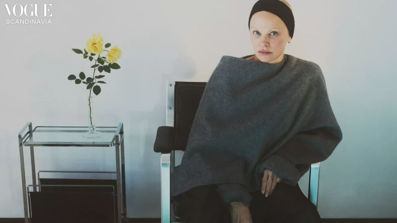 Portrait of adult female model with headband, sitting in chair with grey wrap; yellow roses in vase on metal and glass cart.
