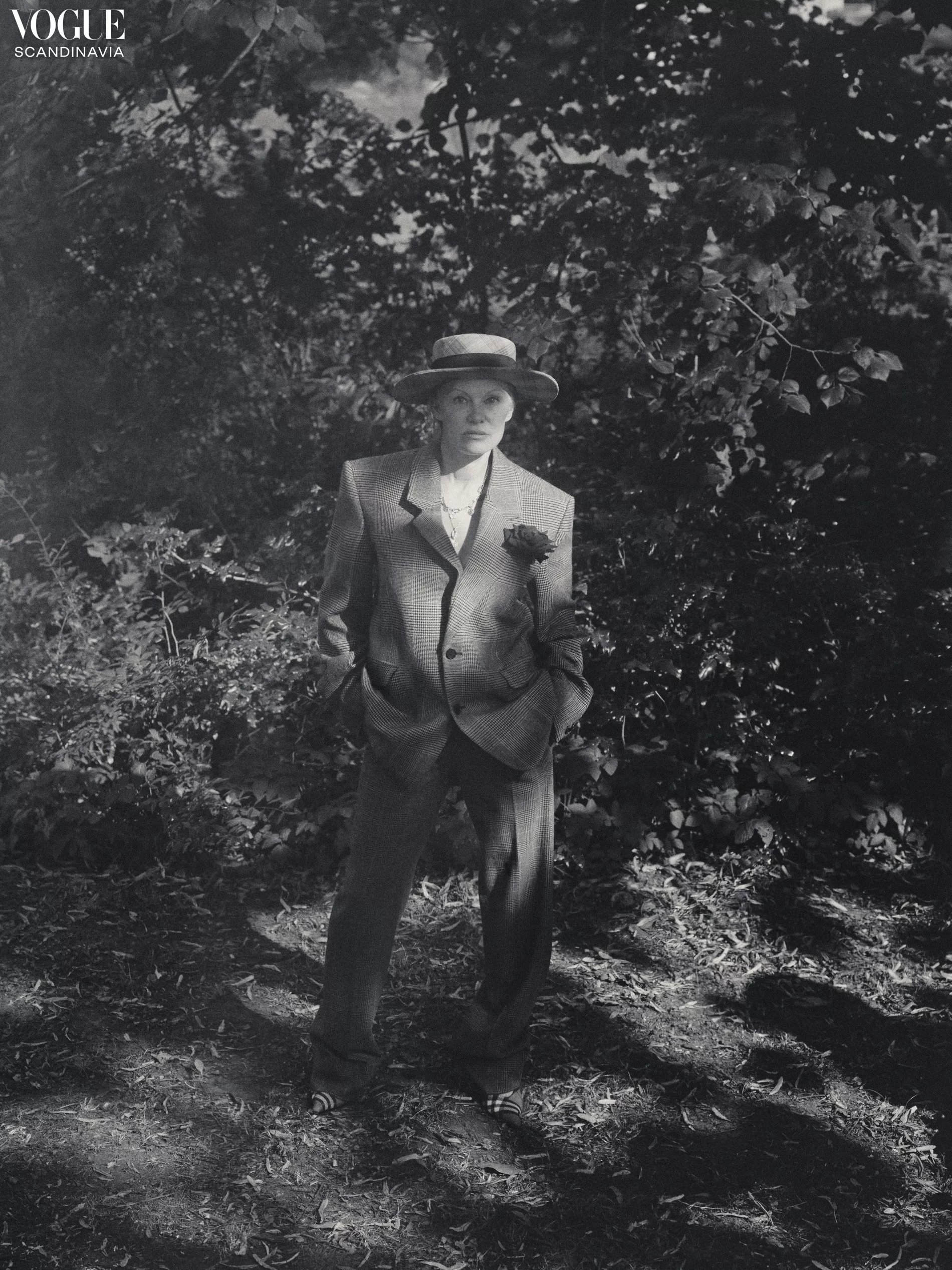 Monochrome fashion portrait of an adult woman in a suit and hat, posing in a wooded area with dappled sunlight, evoking a vintage style.