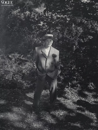 Monochrome fashion portrait of an adult woman in a suit and hat, posing in a wooded area with dappled sunlight, evoking a vintage style.