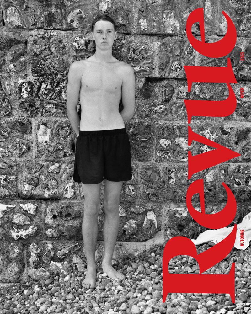 Black and white medium shot of a topless slender male model standing on a pebble beach in front of a stone wall, magazine cover.