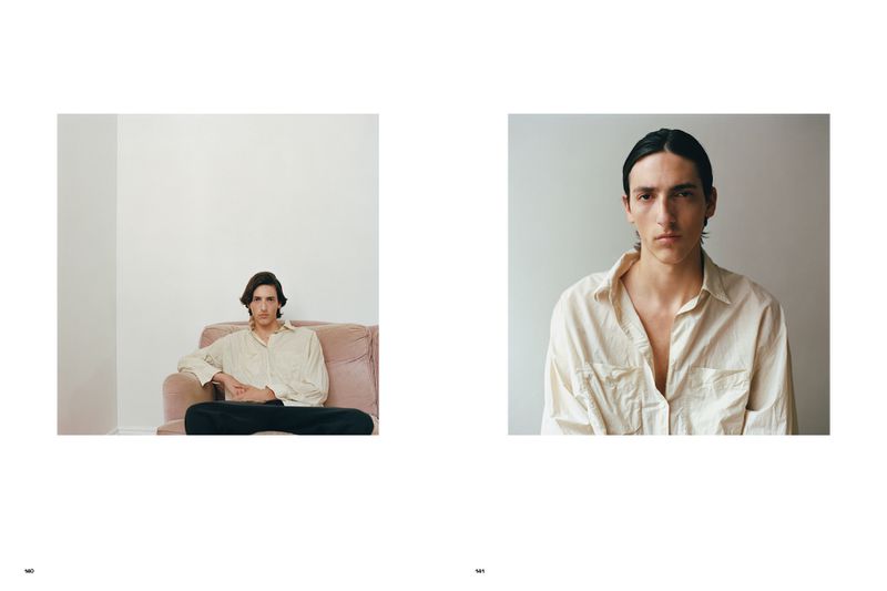 Portrait of a young adult male sitting on a pink couch in front of a white wall. He is wearing an unbuttoned cream-colored shirt.