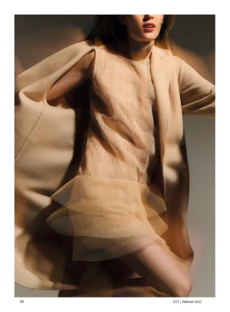 Model wearing a beige sleeveless textured dress and matching coat in motion against a dark studio background