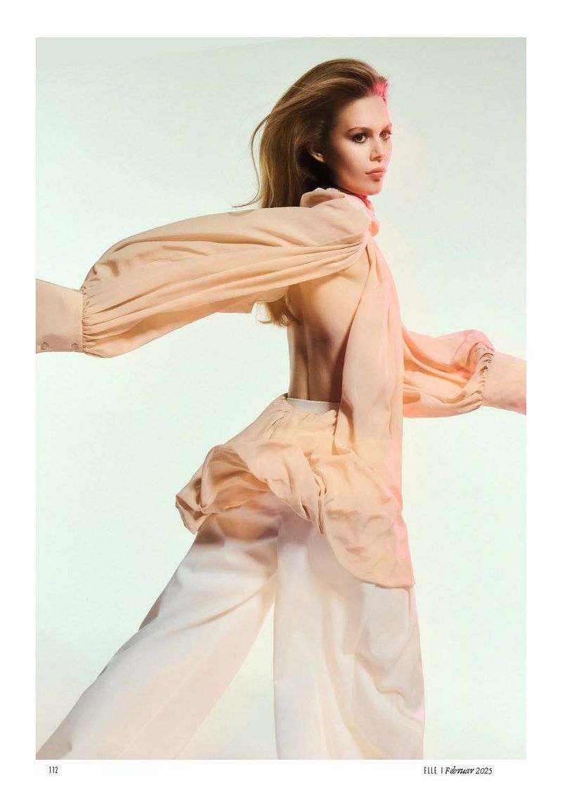 Fashion editorial shot of a blonde woman wearing a loose peach top and white pants, posing in a photo studio with bright lighting.