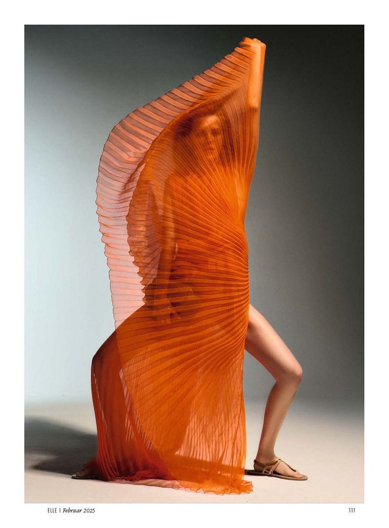 Fashion editorial shot of a woman posing in front of a grey background, draped in a pleated orange fabric.