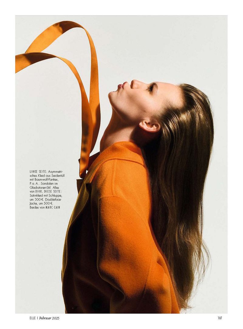 Woman in orange jacket looks up at flowing orange ribbons in a fashion editorial photo against a bright white background.
