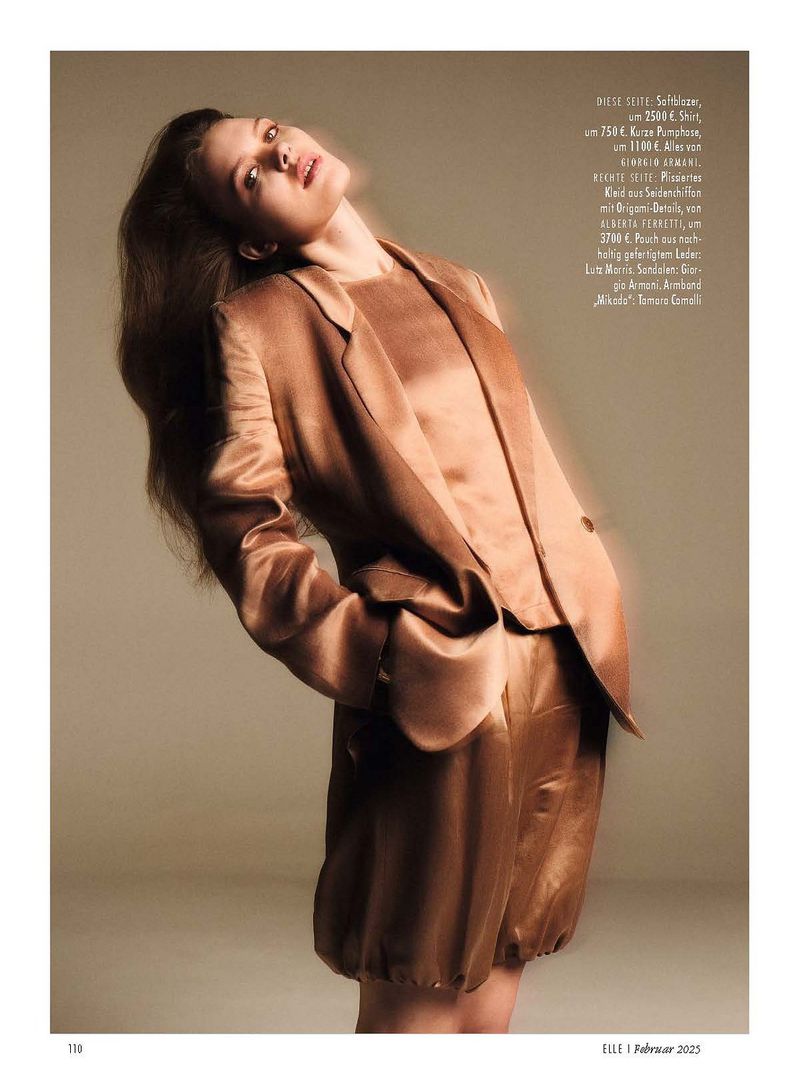 Brunette woman poses in a silky gold-brown blazer and matching shorts, shot from a low angle against a neutral backdrop.