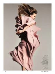 Model in a fluid pink pleated dress captured mid-motion with flying hair against a neutral grey studio background for Elle Germany.