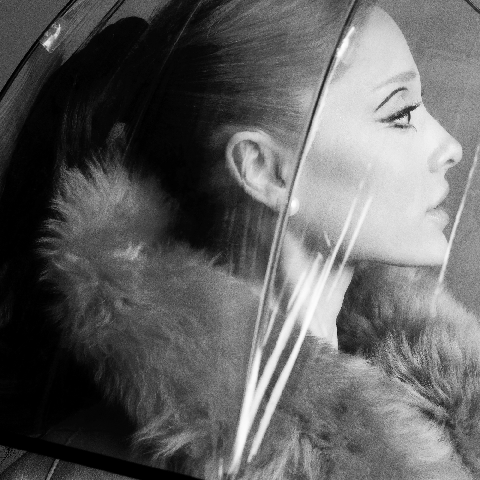 Monochrome portrait of a woman in a fur coat under a clear umbrella, featuring detailed eye makeup and a subtle expression.