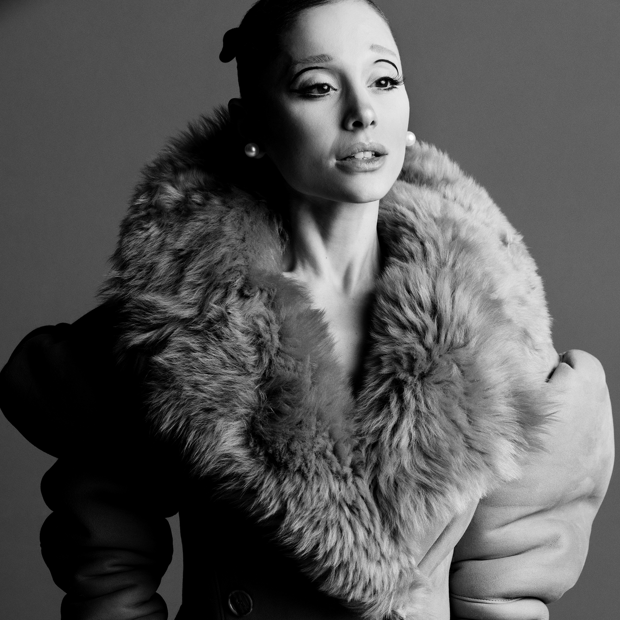 Black and white portrait of an adult woman wearing a fur coat, looking up and away, in a photo studio setting with even lighting.