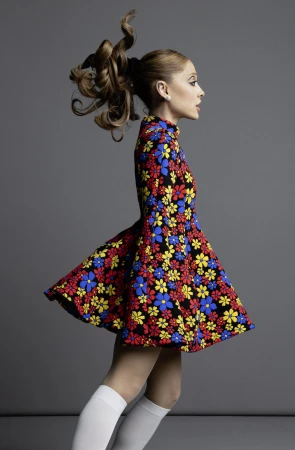 Profile shot of a woman in a 1960s-style floral mini-dress with her hair in a high ponytail, posing against a gray studio backdrop.