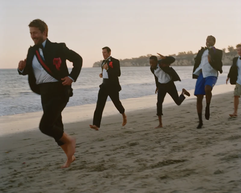 A group of men in tuxedos running barefoot on a sandy beach at sunset, with trees and the ocean visible in the soft-focus background.