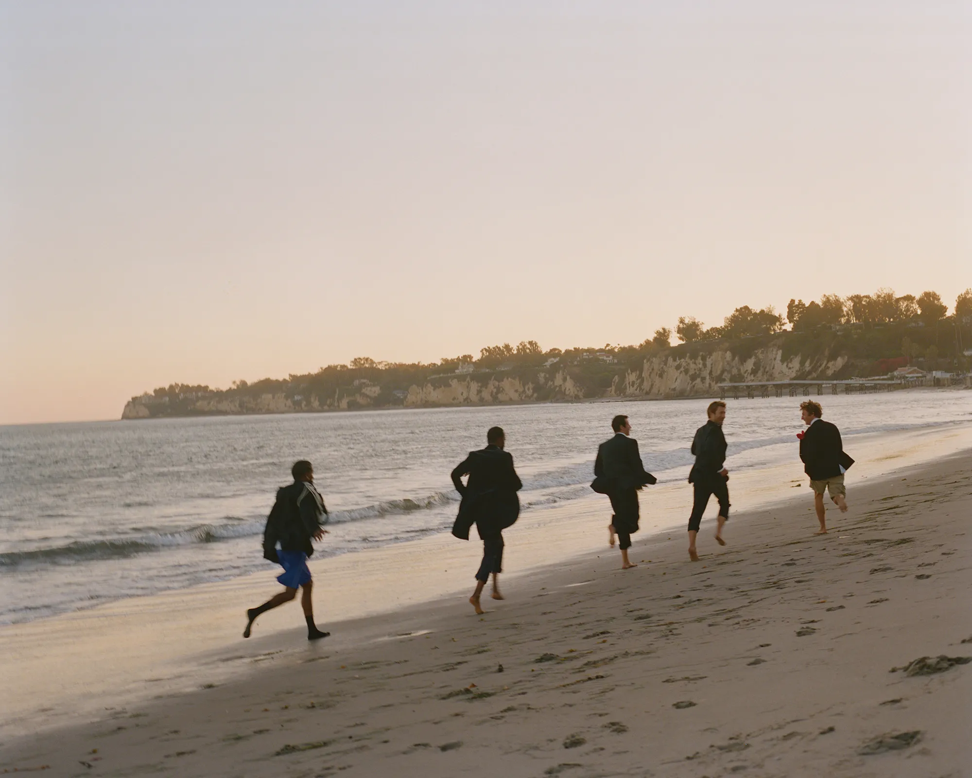 Five people run barefoot on a sandy beach along the ocean shoreline under a bright sky with distant bluffs and green vegetation.