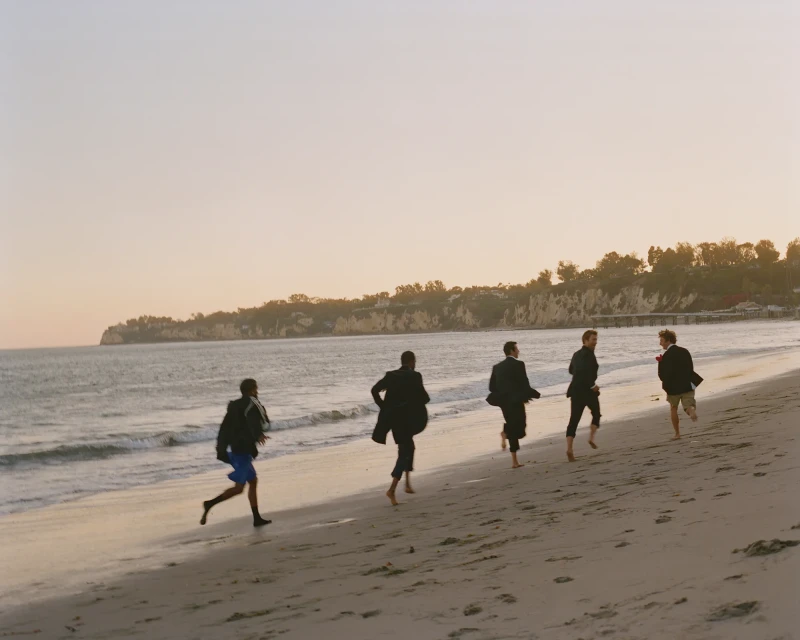 Five people run barefoot on a sandy beach along the ocean shoreline under a bright sky with distant bluffs and green vegetation.