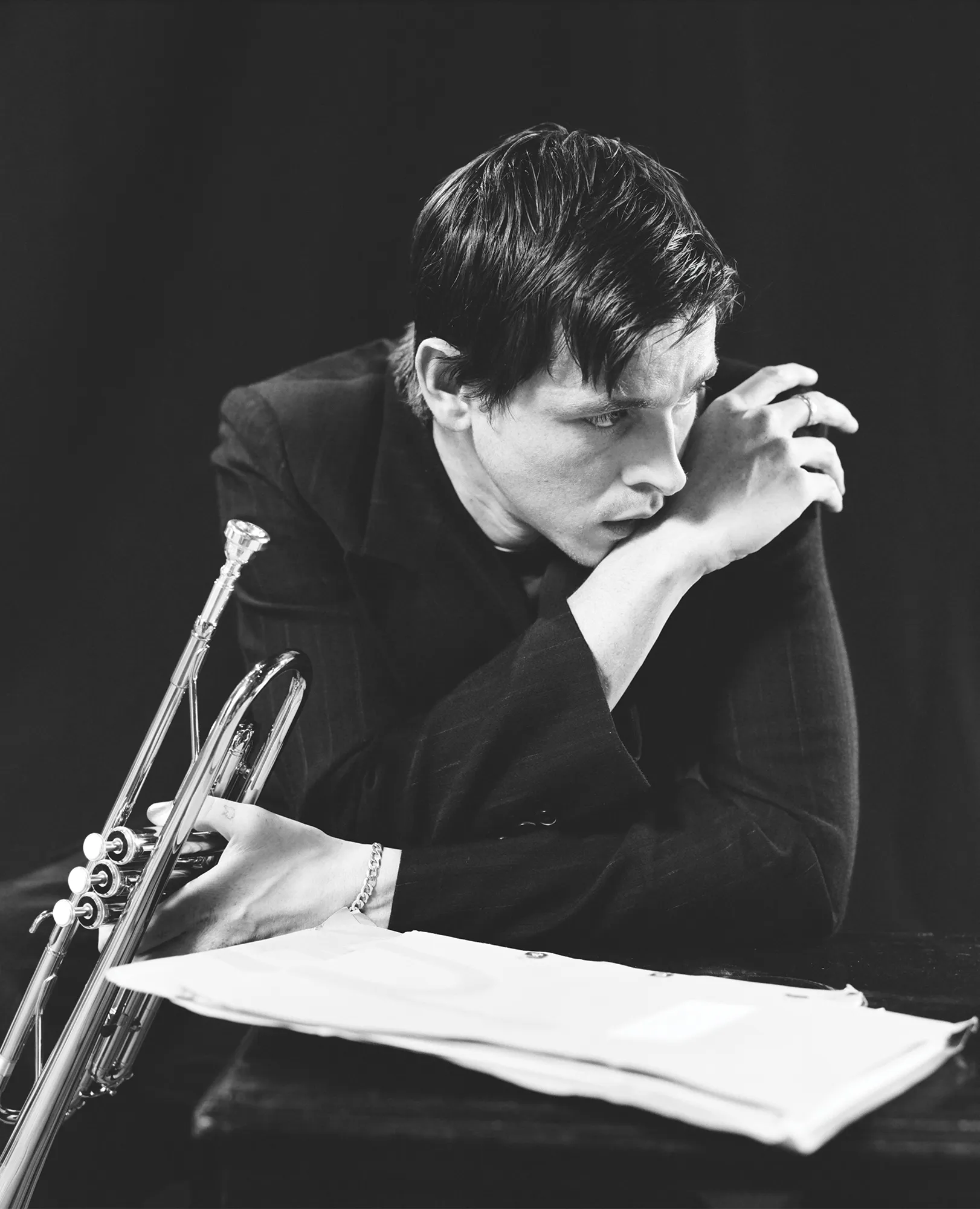 Black and white portrait of a thoughtful musician holding a trumpet in a photo studio. Sheet music and a dark backdrop complete the scene.