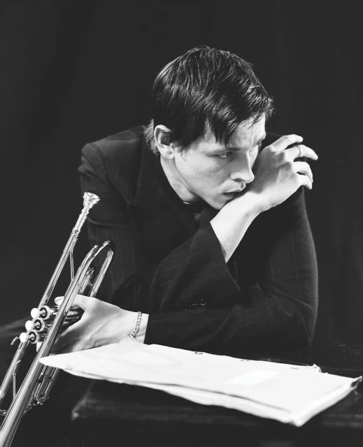 Black and white portrait of a thoughtful musician holding a trumpet in a photo studio. Sheet music and a dark backdrop complete the scene.