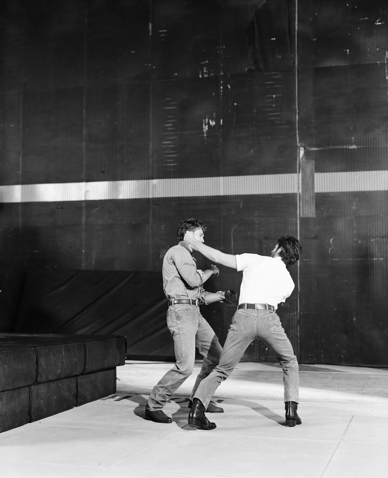 Two men in an indoor studio grappling and fighting in a theatrical performance with dramatic lighting, wearing jeans and boots.