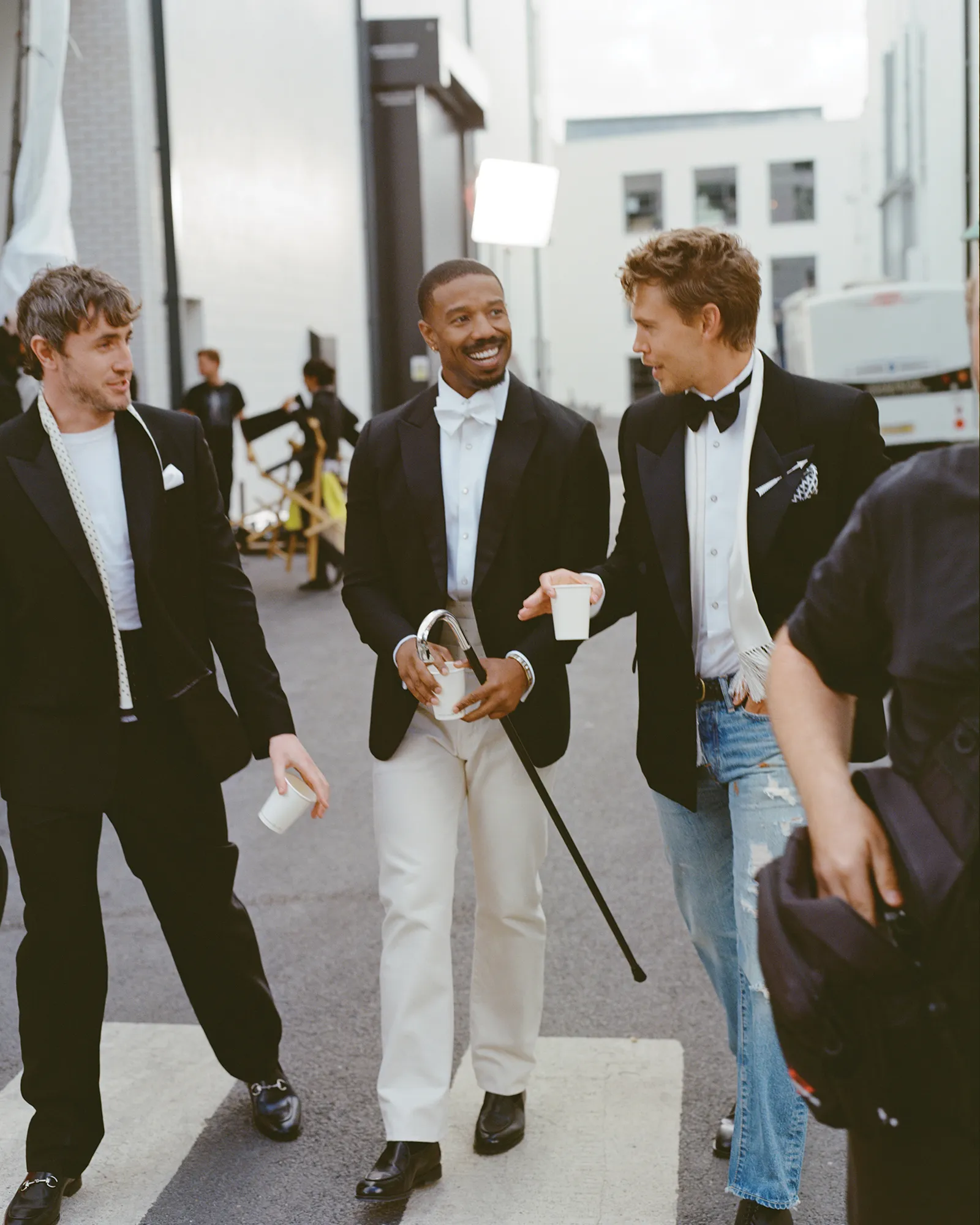 Three men in tuxedos walk on a zebra crossing outdoors. Michael B. Jordan holds a cane. Austin Butler wears jeans with his tuxedo.