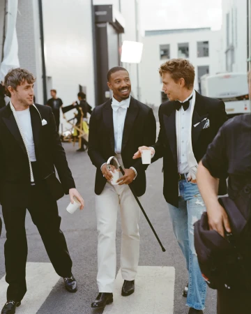 Three men in tuxedos walk on a zebra crossing outdoors. Michael B. Jordan holds a cane. Austin Butler wears jeans with his tuxedo.
