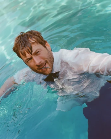 Adult man in a white shirt and bow tie is submerged in bright blue water. Wet hair is slicked back on the fair-skinned male model.