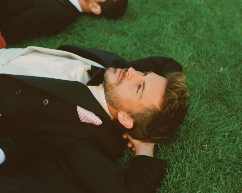 Two men in formal wear are lying on green grass outside, one in the foreground, with focus on his face and upper body.