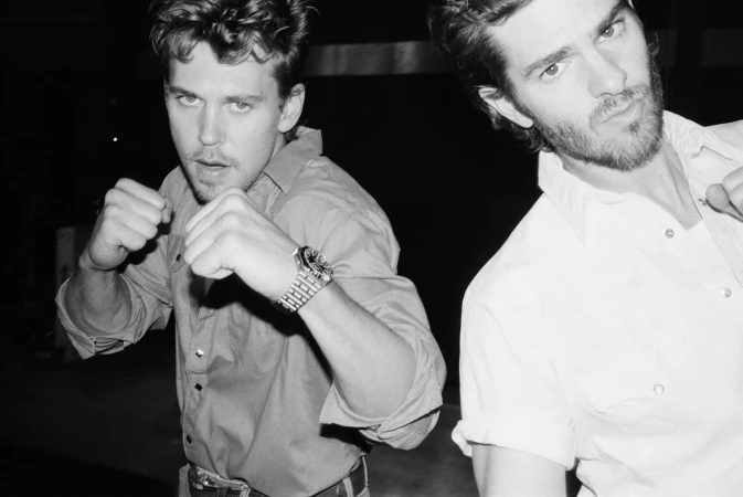 Black-and-white portrait of two young adult males posing playfully, one with fists up, set against a dark, nondescript background.