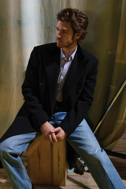 A man wearing a black coat and blue jeans sits on a box labeled Matthews against a neutral backdrop in a studio setting.
