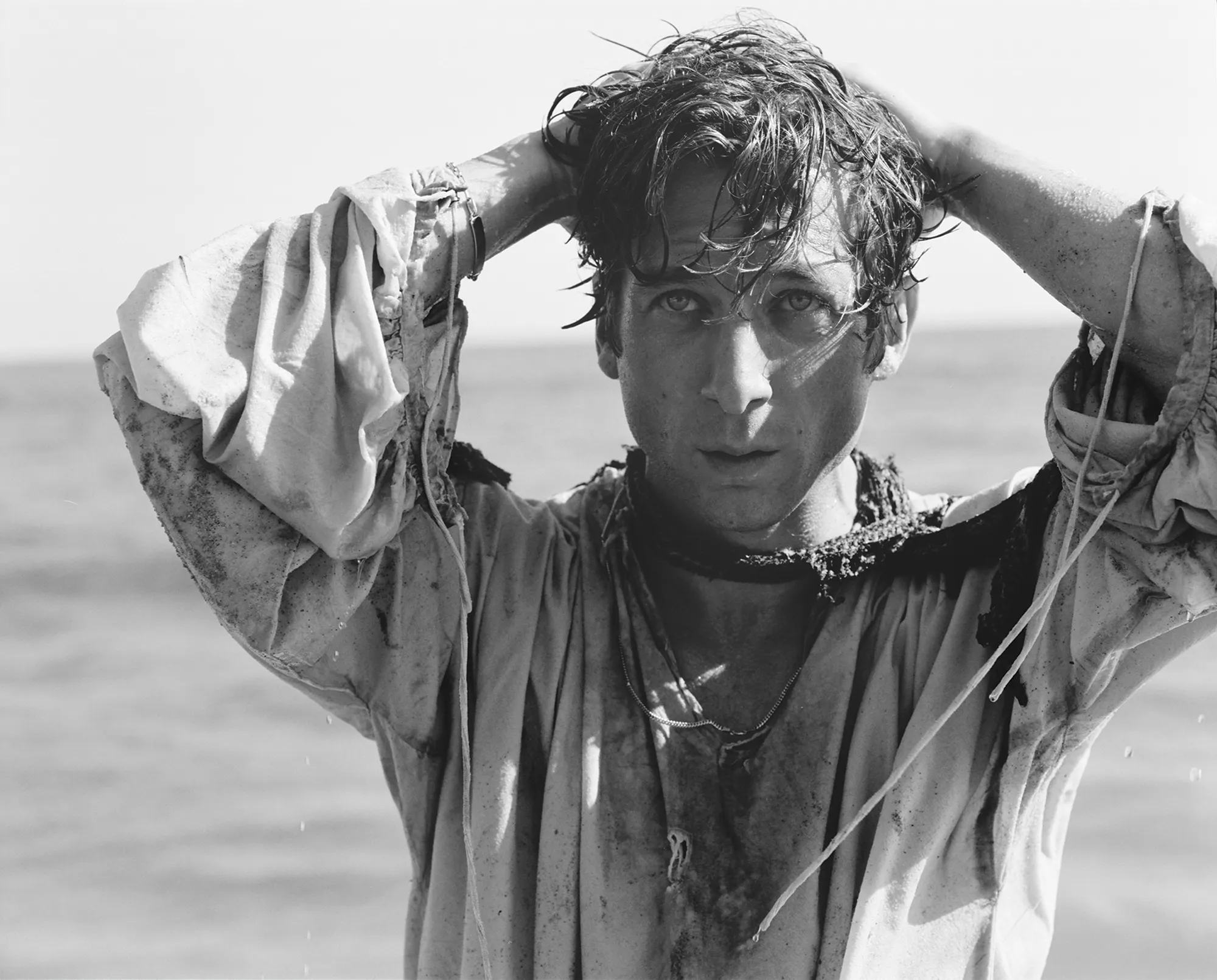 Monochrome portrait of a man at the beach with wet hair and disheveled clothing, his hands atop his head against the ocean backdrop.